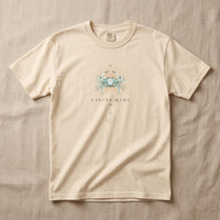 Ivory Cancer Mama graphic tee laid flat on neutral linen surface, featuring celestial crab artwork and gold sunburst design.