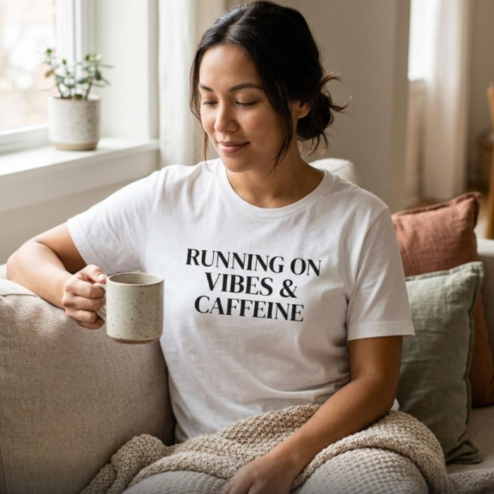 Woman wearing white “Running on Vibes & Caffeine” graphic t-shirt sitting with coffee, cozy lifestyle outfit