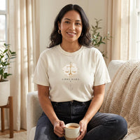 Ivory Libra Mama graphic t-shirt worn by a Latina woman seated indoors in soft natural light, featuring gold scale design with celestial details.