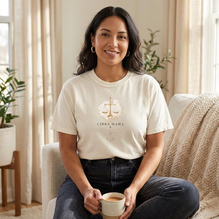 Ivory Libra Mama graphic t-shirt worn by a Latina woman seated indoors in soft natural light, featuring gold scale design with celestial details.