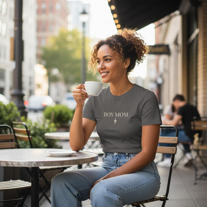 Woman sitting outdoors holding a cup, wearing a 'BOY MOM' t-shirt.