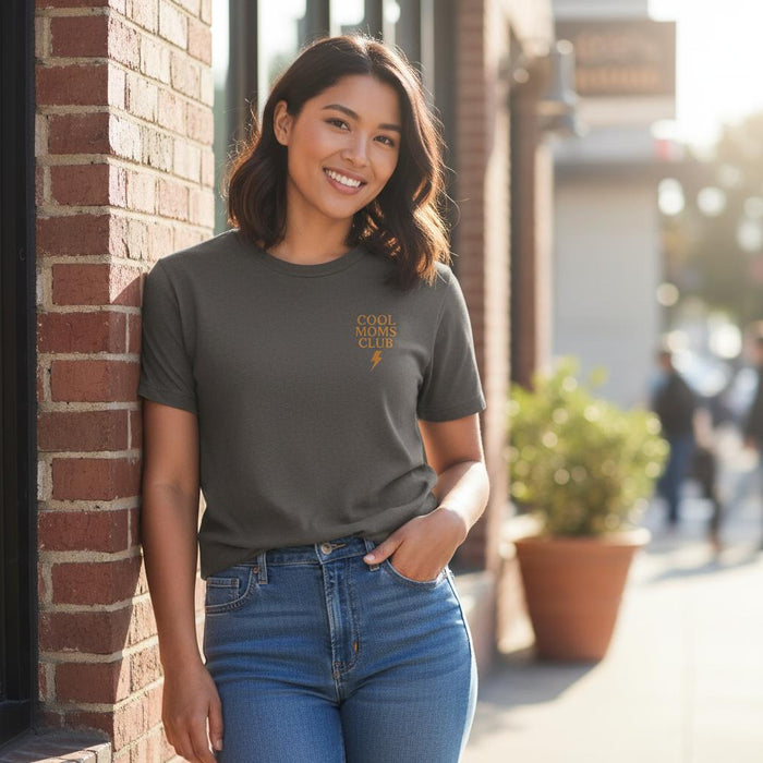 Woman wearing a Comfort Colors t-shirt in Pepper with 'Cool Moms Club' text, standing against a brick wall.