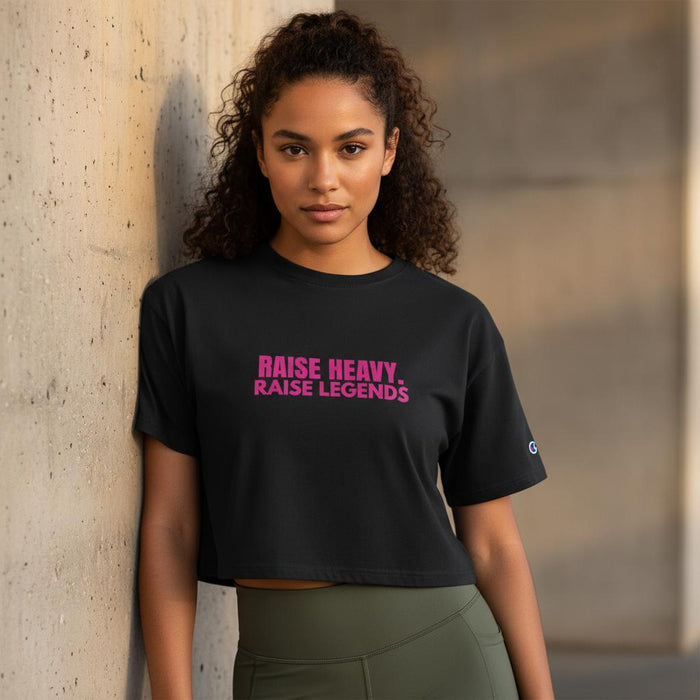 Woman wearing a black cropped Champion tee with pink “Raise Heavy. Raise Legends.” graphic leaning against a wall