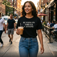 Woman wearing black “Running on Vibes & Caffeine” graphic t-shirt walking in city with iced coffee, casual everyday outfit