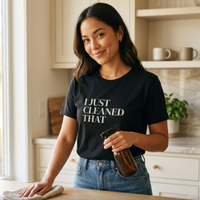 Woman wearing black “I Just Cleaned That” t-shirt holding cleaning spray, relatable mom life moment
