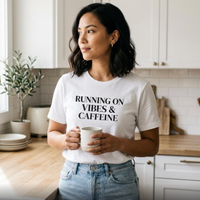 Woman wearing white “Running on Vibes & Caffeine” t-shirt in kitchen holding mug, casual everyday style