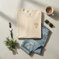 Folded beige t-shirt with 'Cool Moms Club' text, jeans, watch, coffee, and sunglasses on a light surface.