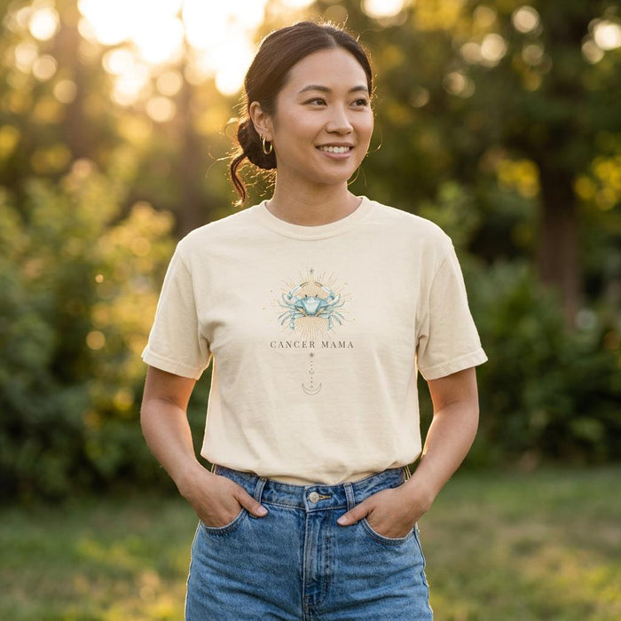 Asian woman wearing an Ivory Cancer Mama graphic t-shirt tucked into blue jeans outdoors in golden hour sunlight with greenery background.
