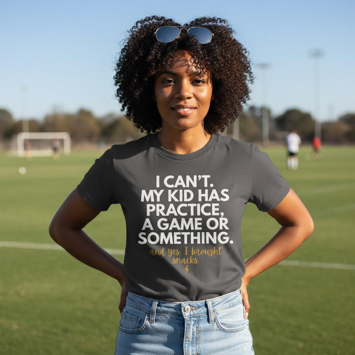 Black woman wearing Pepper graphic tee that reads “I Can’t. My Kid Has Practice. And yes, I brought snacks.” standing confidently on a soccer field with hands on hips, styled with light denim jeans in natural sunlight.