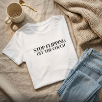 White “Stop Flipping Off the Couch” graphic t-shirt flat lay styled with jeans and cozy neutral accessories