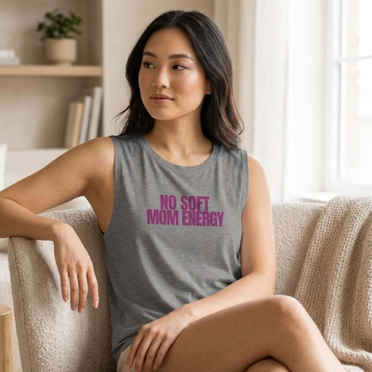 Woman sitting on couch wearing gray “No Soft Mom Energy” tank top in relaxed setting