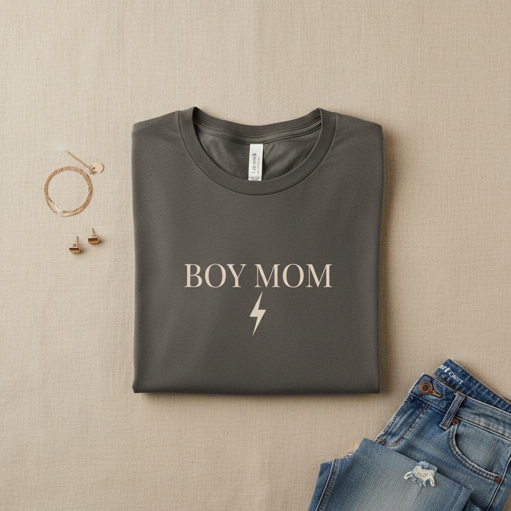 Folded gray t-shirt with 'BOY MOM' text on a beige surface with jeans and jewelry.