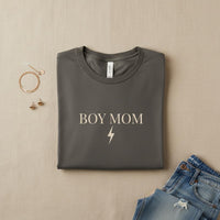 Folded gray t-shirt with 'BOY MOM' text on a beige surface with jeans and jewelry.