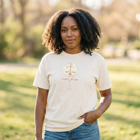 Ivory Libra Mama zodiac t-shirt worn by a woman of color outdoors in warm sunlight, one hand in pocket, gold scale artwork centered on chest.