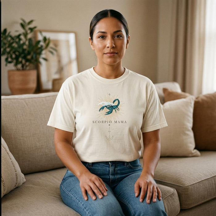 Latina woman wearing Scorpio Mama ivory zodiac graphic t-shirt seated indoors on neutral sofa
