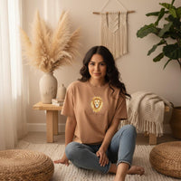 Load image into Gallery viewer, Latina woman sitting indoors wearing a sandstone Leo Mama zodiac t-shirt with celestial lion design in a cozy neutral living room