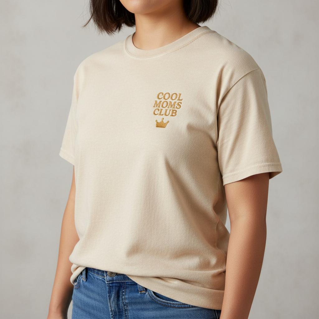 Female model wearing an ivory t-shirt with 'Cool Moms Club' text and a crown graphic on a plain background