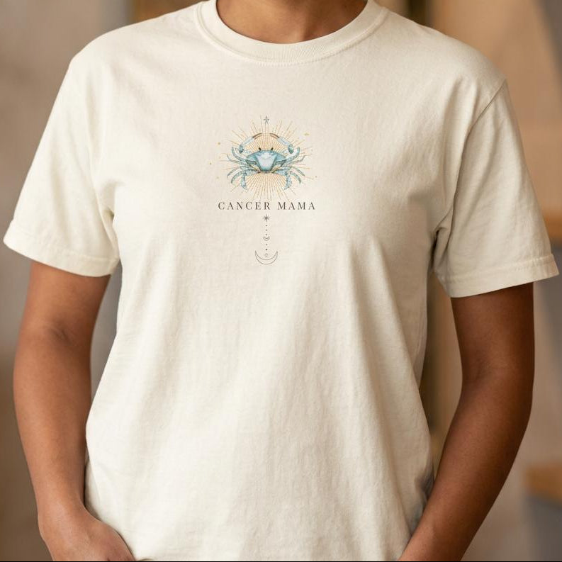 Close-up detail of Ivory Cancer Mama zodiac t-shirt showing watercolor crab chest design with gold sunburst rays and vertical moon phase accents.