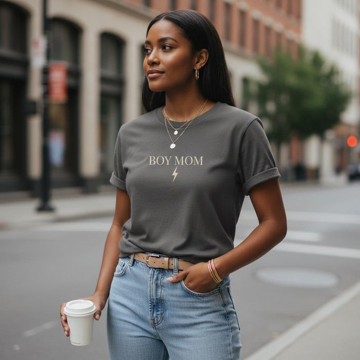 Woman wearing a gray t-shirt with 'BOY MOM' text, holding a coffee cup on a city street.