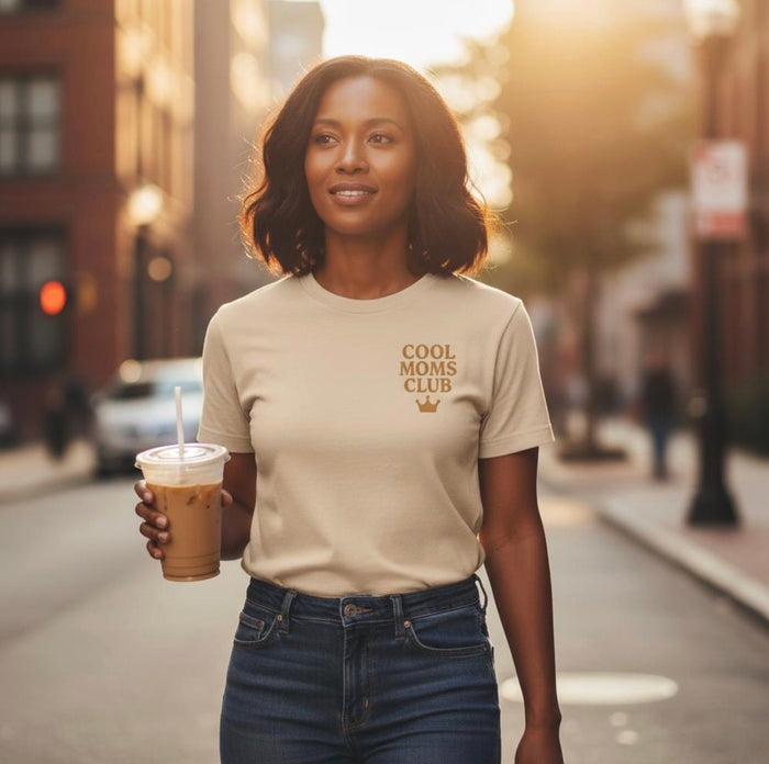Woman wearing a Comfort Colors t-shirt in Ivory with 'Cool Moms Club' text and crown icon holding a drink on a city street.