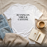 White “Running on Vibes & Caffeine” graphic t-shirt flat lay with jeans and coffee, neutral styled outfit