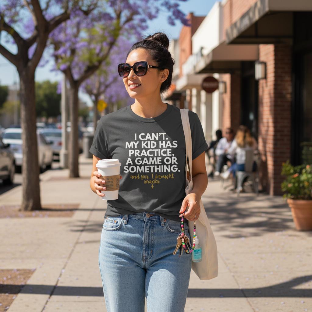 Asian woman wearing Pepper “I Can’t My Kid Has Practice” shirt tucked into jeans, walking on a sunny sidewalk holding coffee and tote bag.