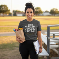 Latina woman wearing Pepper snack mom graphic tee holding a paper bag filled with snacks and a clipboard while standing near bleachers at a sports field.