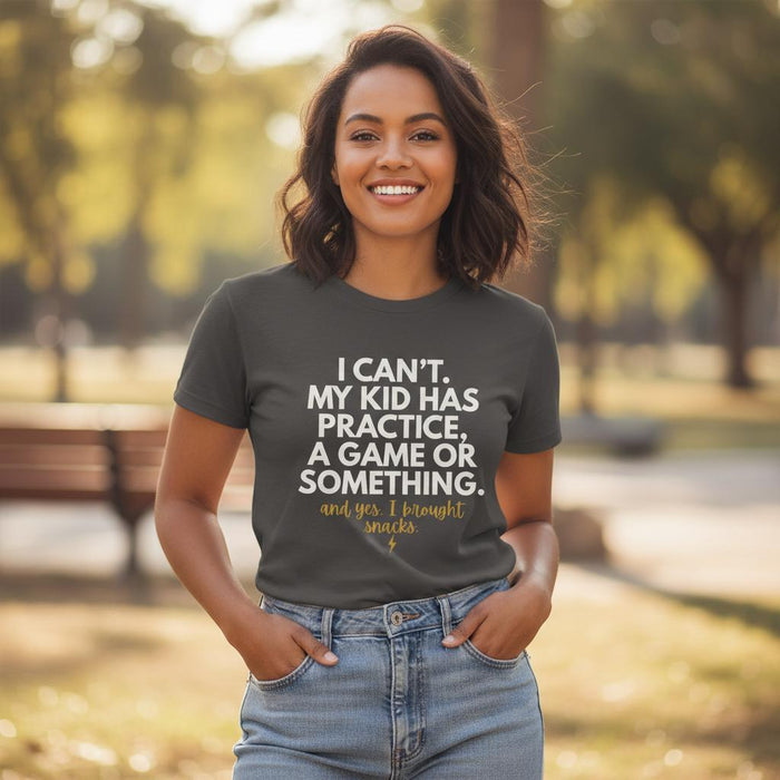 Woman wearing Pepper “I Can’t My Kid Has Practice” graphic tee tucked into high-waisted jeans, smiling outdoors in warm golden hour light at a park.